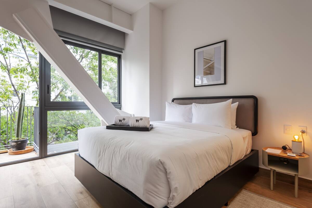 Un dormitorio moderno con una cama grande, una mesita de noche, una lámpara de noche, un cuadro enmarcado en la pared y vistas a árboles a través de un ventanal amplio.