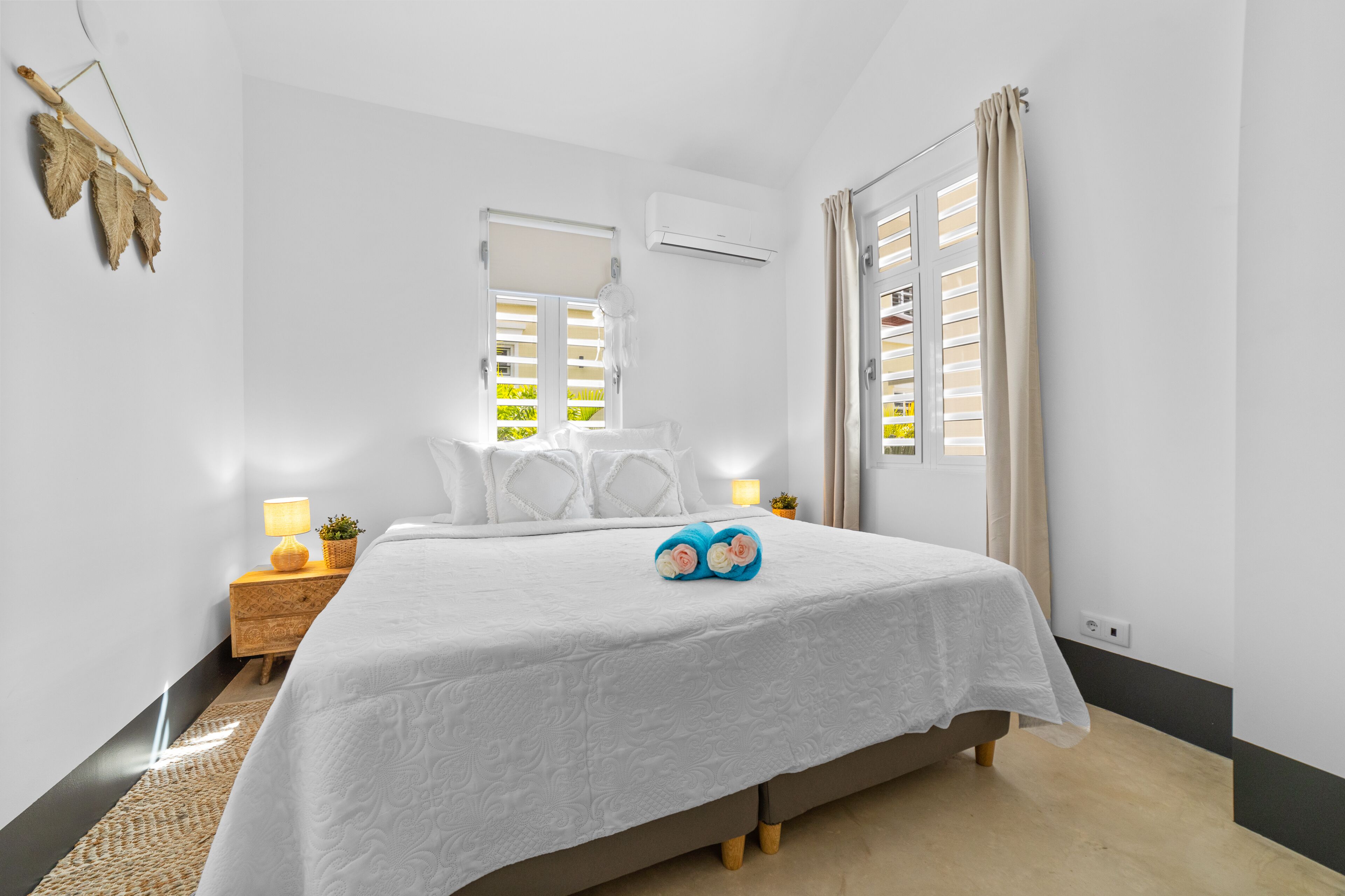 A bedroom with a bed, bedside lamps, a small table with a lamp, a potted plant, and a window with blinds.