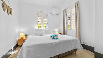 A bedroom with a bed, bedside lamps, a small table with a lamp, a potted plant, and a window with blinds.