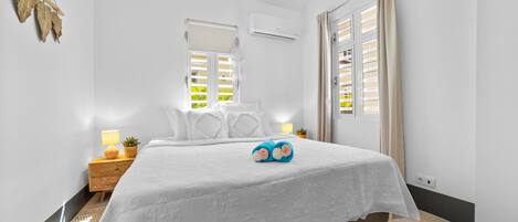 A bedroom with a bed, bedside lamps, a small table with a lamp, a potted plant, and a window with blinds.