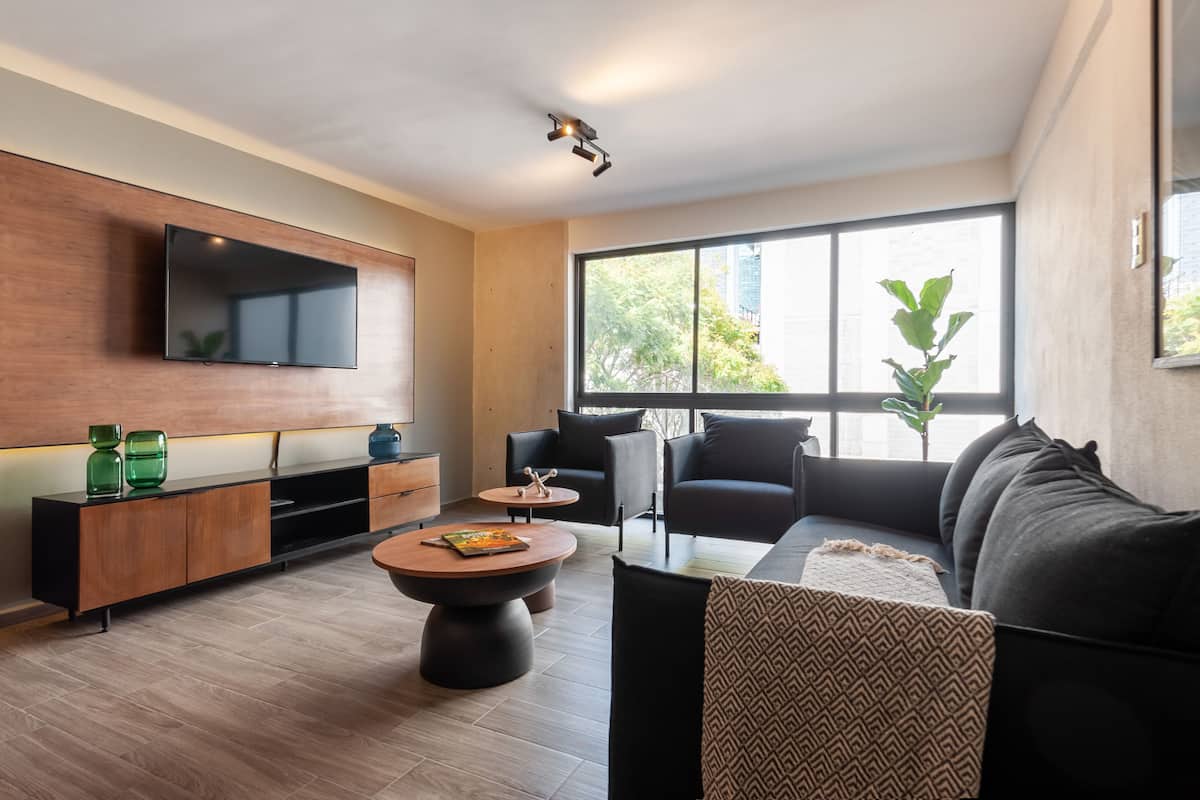 Una sala moderna con un televisor de pantalla plana montado en la pared, un mueble de madera para entretenimiento, un sofá negro, una mesa de centro redonda y un ventanal con vista a árboles.