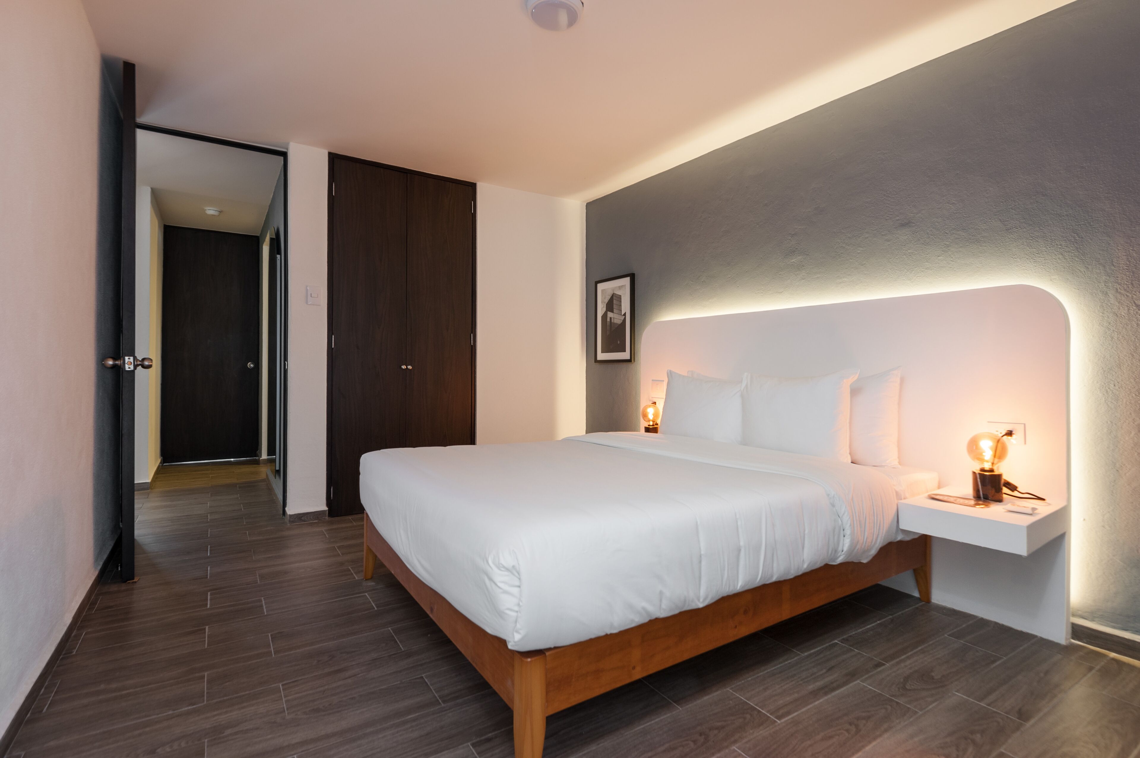 A modern bedroom with a wooden bed, white linens, a bedside table with a lamp, and a framed picture on the wall.