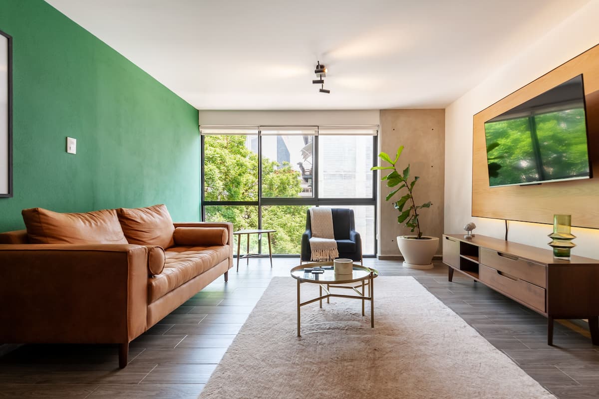 Una sala de estar moderna con un sofá de cuero marrón, una mesa de centro redonda, un sillón negro y un ventanal con vistas a la ciudad.