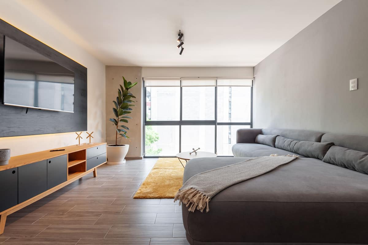Una sala de estar moderna con un sofá grande de color gris, un mueble bajo de televisión de madera, una planta en maceta y un ventanal que proporciona luz natural.