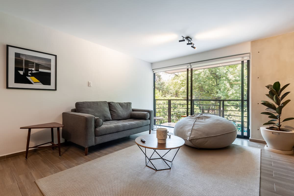 Una sala de estar moderna con un sofá gris, una mesita redonda, un sillón de tina y un ventanal con vistas a zonas verdes.