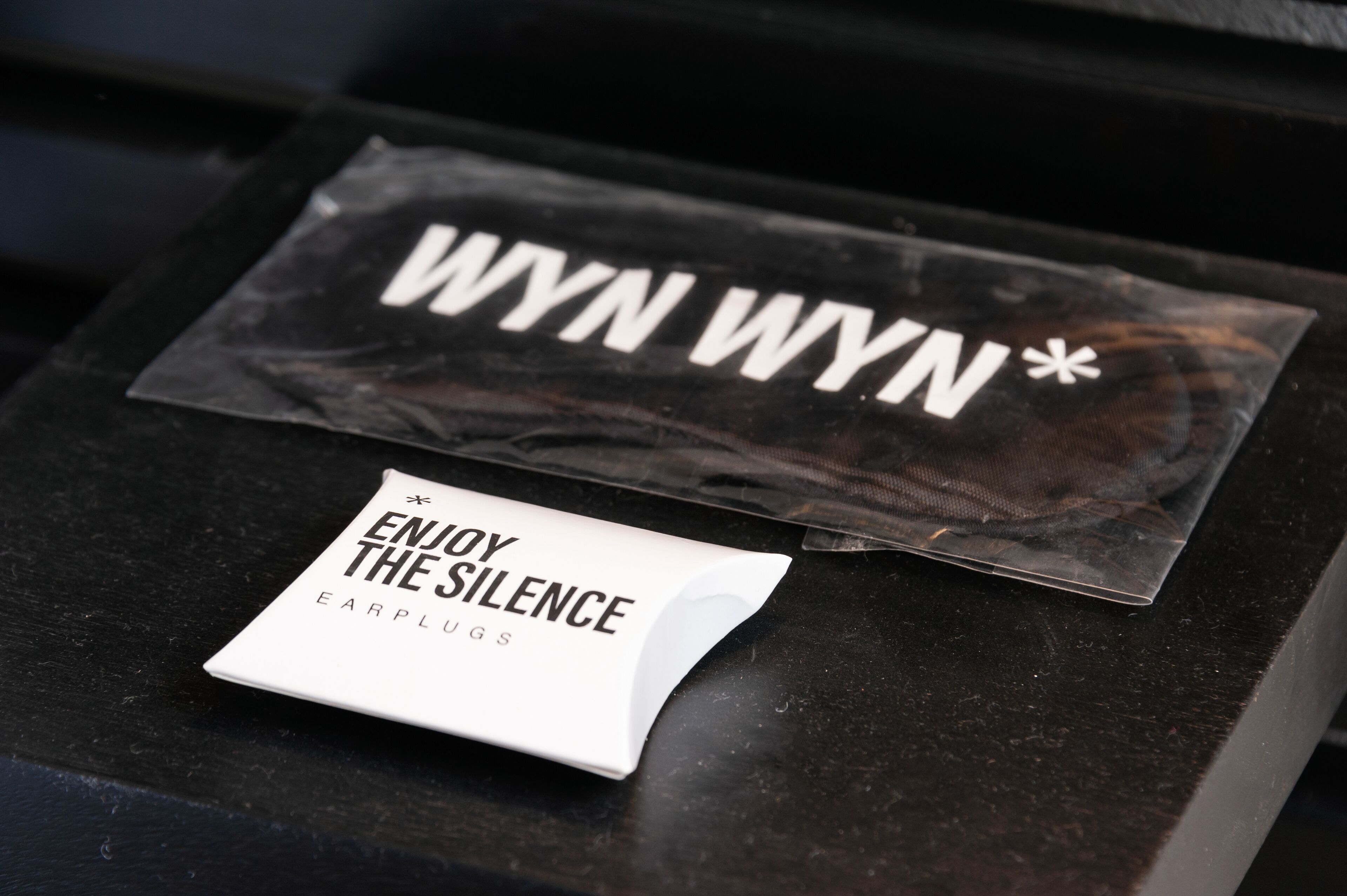 A black surface with a white box labeled "ENJOY THE SILENCE EARPLUGS" and a plastic bag with "WYNN WYNN" text.
