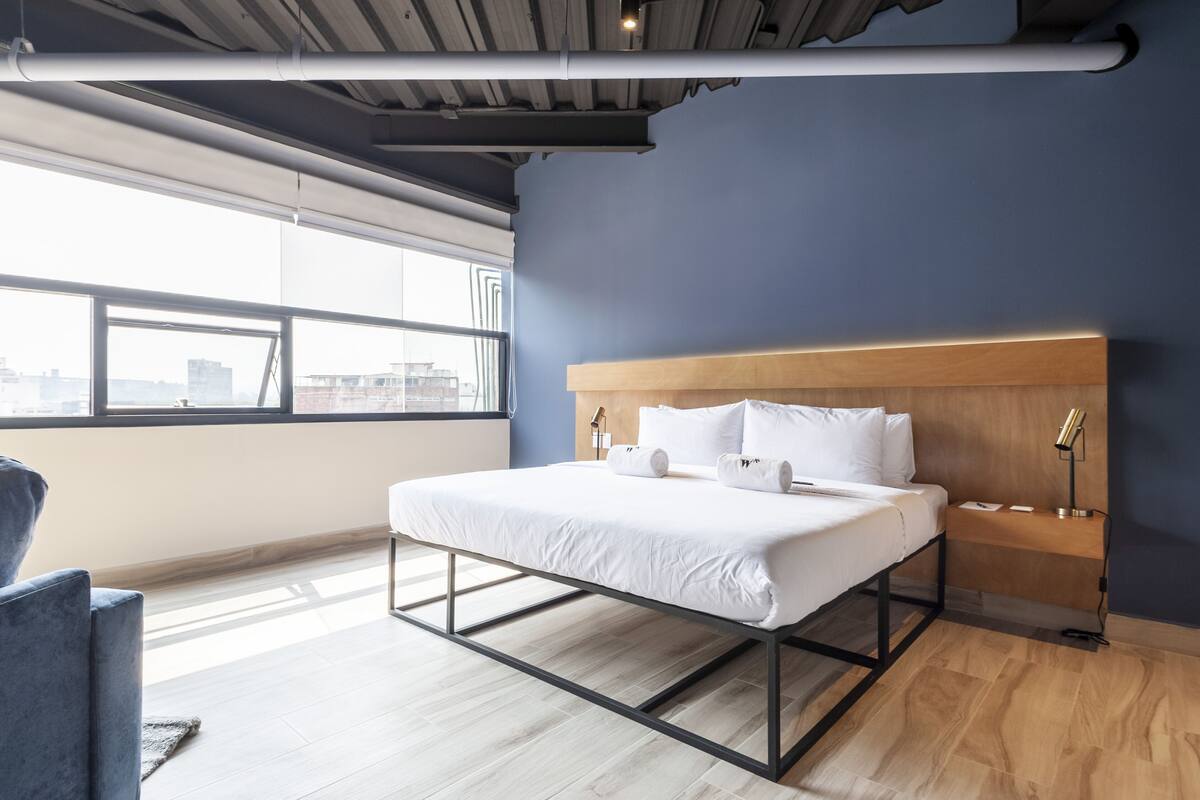 Une chambre d’hôtel moderne, équipée d’un lit, d’une table de chevet, d’une lampe et offrant une vue sur la ville grâce à de grandes fenêtres.