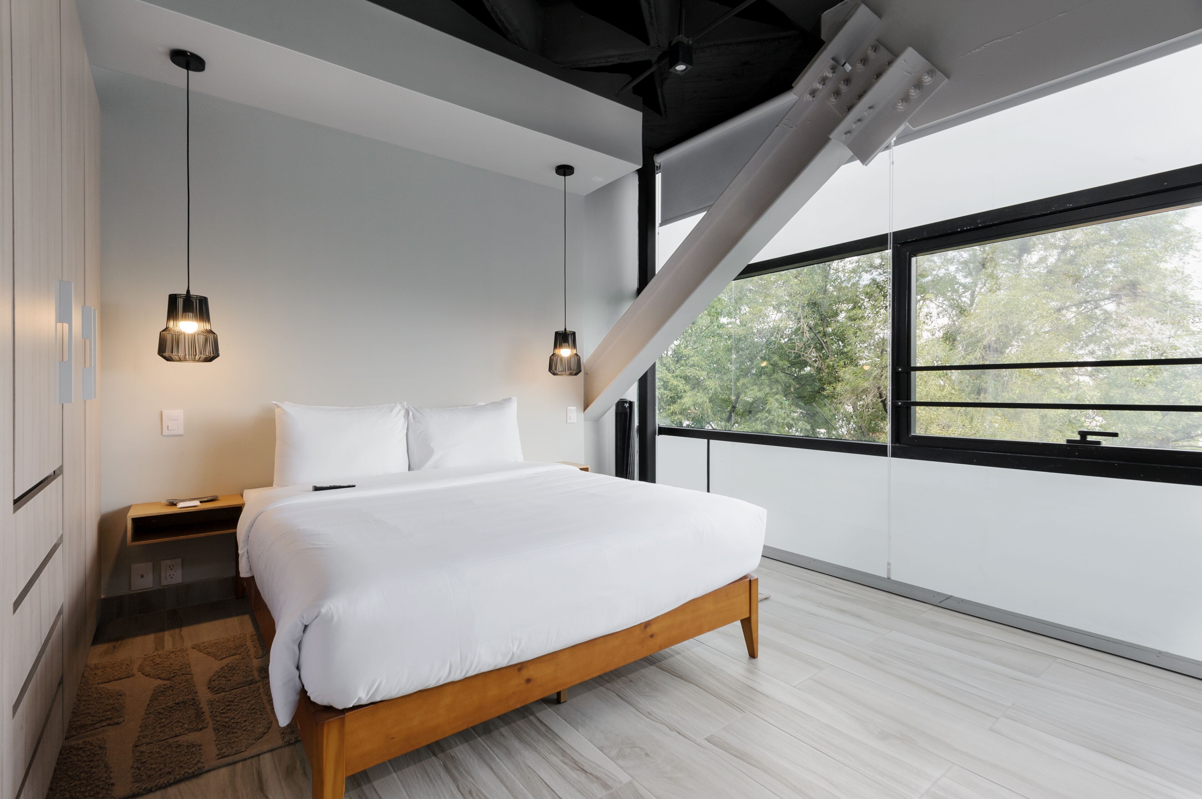 Une chambre moderne avec un grand lit, deux lampes suspendues, une table de chevet et une vue sur les arbres à travers une grande fenêtre.