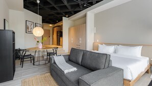 Een moderne hotelkamer met een grijze bank, een bed met witte lakens, een zithoek met een ronde tafel en stoelen, en een keuken met houten kasten.