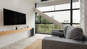 Een moderne woonkamer met een grote flatscreen televisie bevestigd aan een houten plank, een grijze bank en een groot raam met uitzicht op de stad.