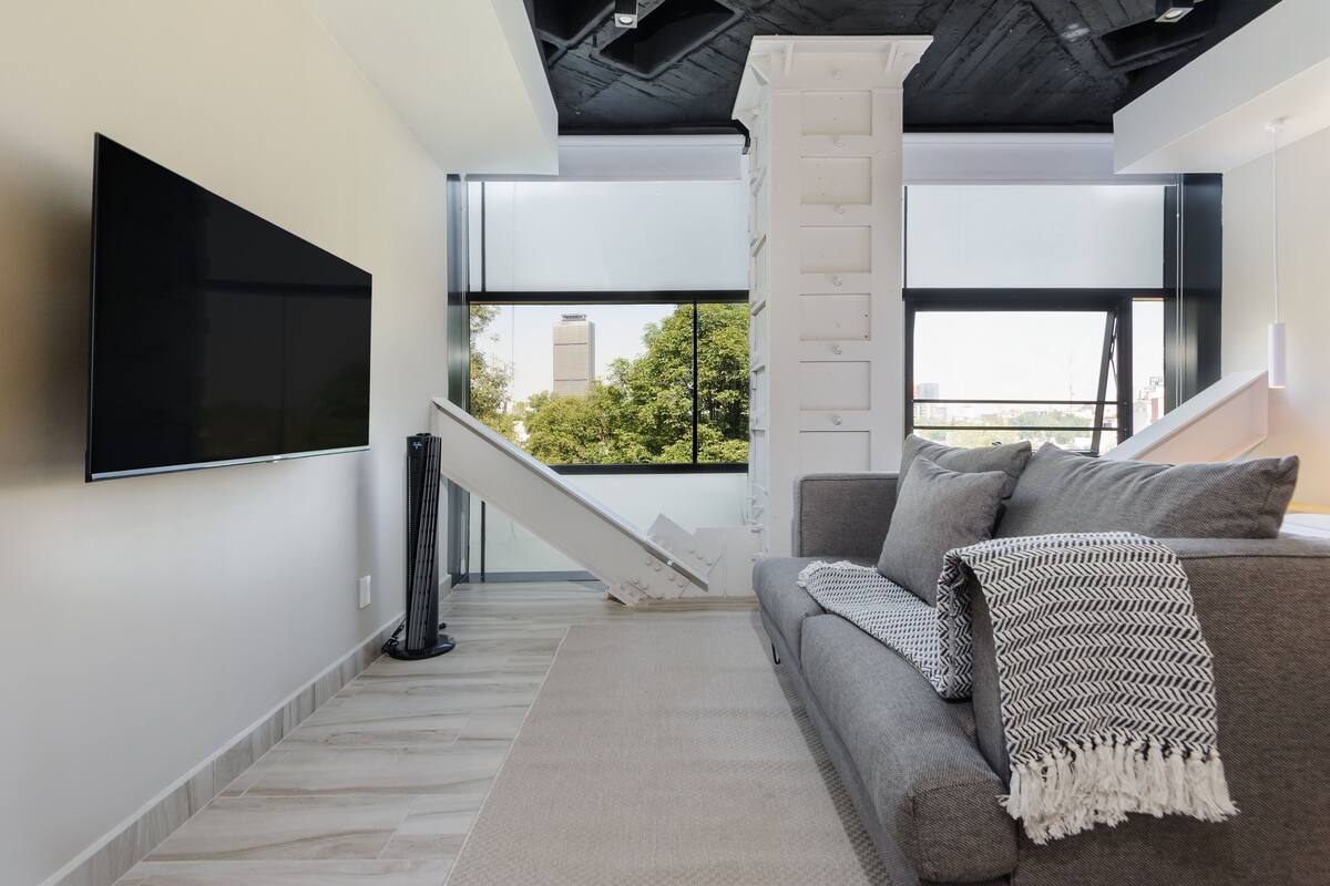 Un salon moderne doté d’un téléviseur à écran plat, d’un canapé gris et d’un escalier menant à un balcon extérieur.