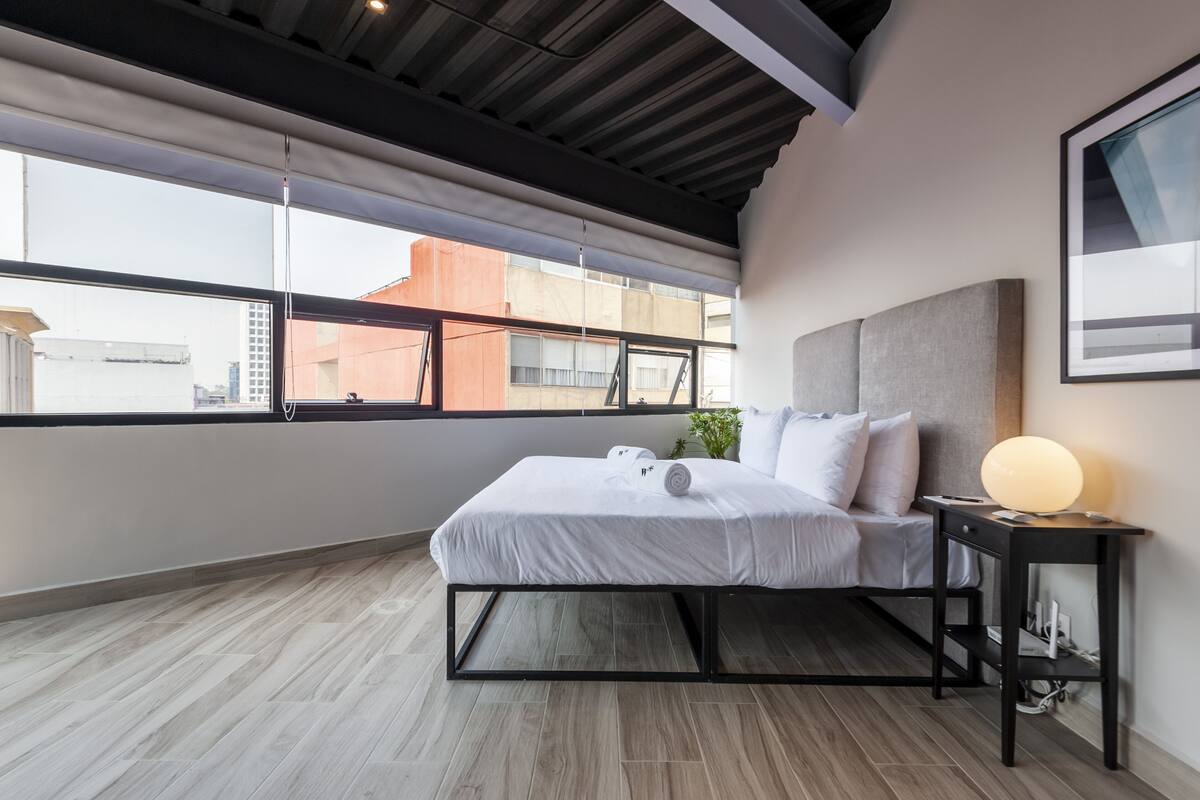 Une chambre d’hôtel moderne avec un grand lit, une table de chevet avec une lampe et une vue sur la ville grâce à de grandes fenêtres.