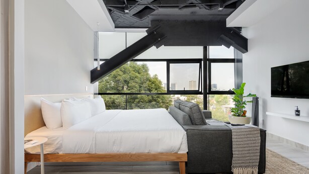 Een moderne slaapkamer met een bed, een bank, een televisie en een groot raam met uitzicht op bomen en gebouwen.