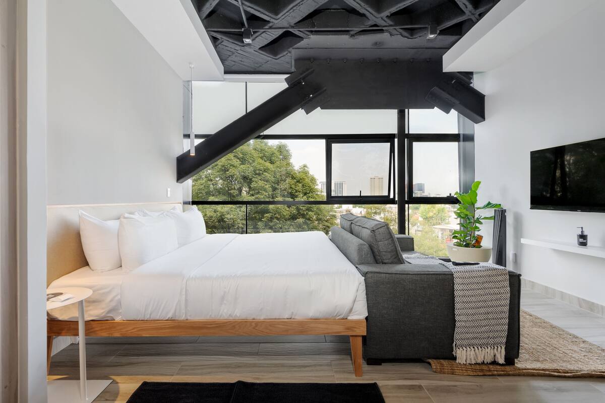 Une chambre moderne avec un lit, un canapé, une télévision et une grande fenêtre donnant sur des arbres et des immeubles.