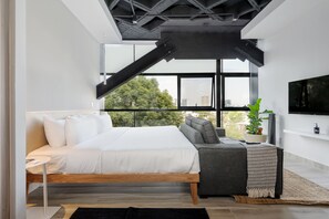 Une chambre moderne avec un lit, un canapé, une télévision et une grande fenêtre donnant sur des arbres et des immeubles.