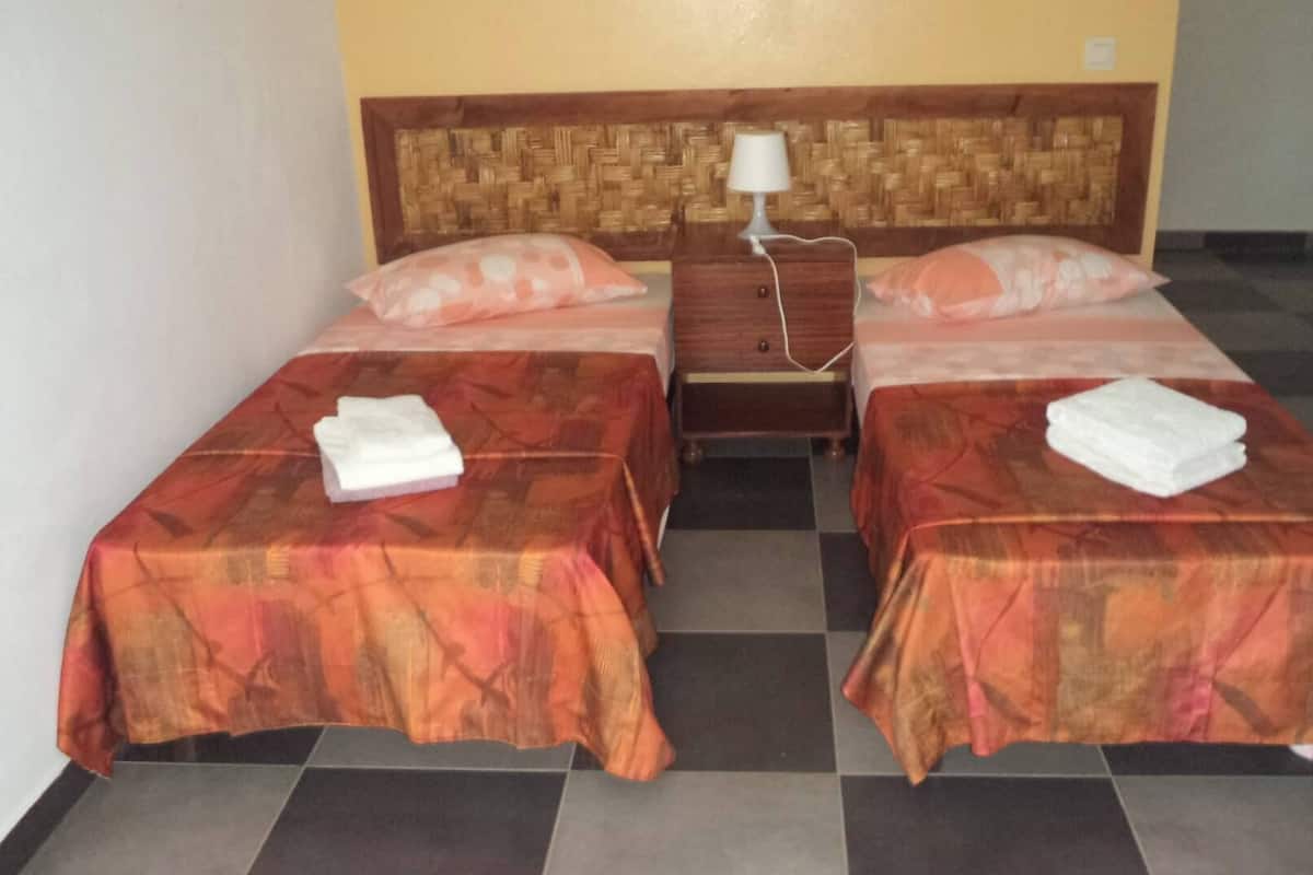 Ein Hotelzimmer mit zwei Einzelbetten, jeweils mit einem Kissen und einem gefalteten Handtuch. Dazwischen steht ein Nachttisch mit einer Lampe.
