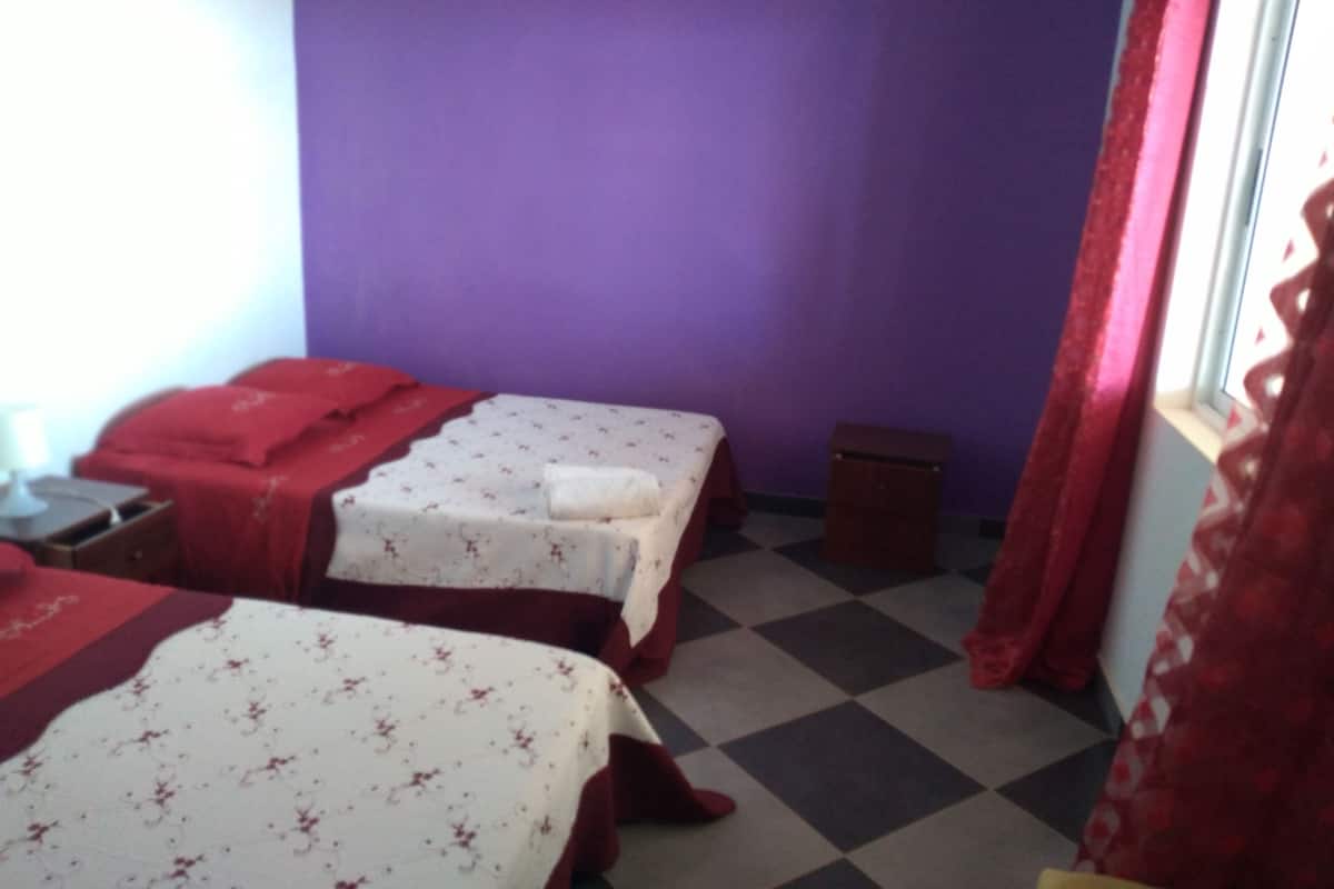 Ein Zimmer mit zwei Betten, violetten Wänden, einem karierten Boden und einem Fenster mit roten Vorhängen.