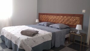 Ein ordentlich gemachtes Bett mit einer gemusterten Bettdecke, zwei Kissen und einem aufgerollten Handtuch an der Seite. Am Kopfteil befindet sich ein Holzpaneel mit einem gewobenen Muster, ein Nachttisch mit einer Lampe und ein Schichtbodenbelag mit Schachbrettmuster.