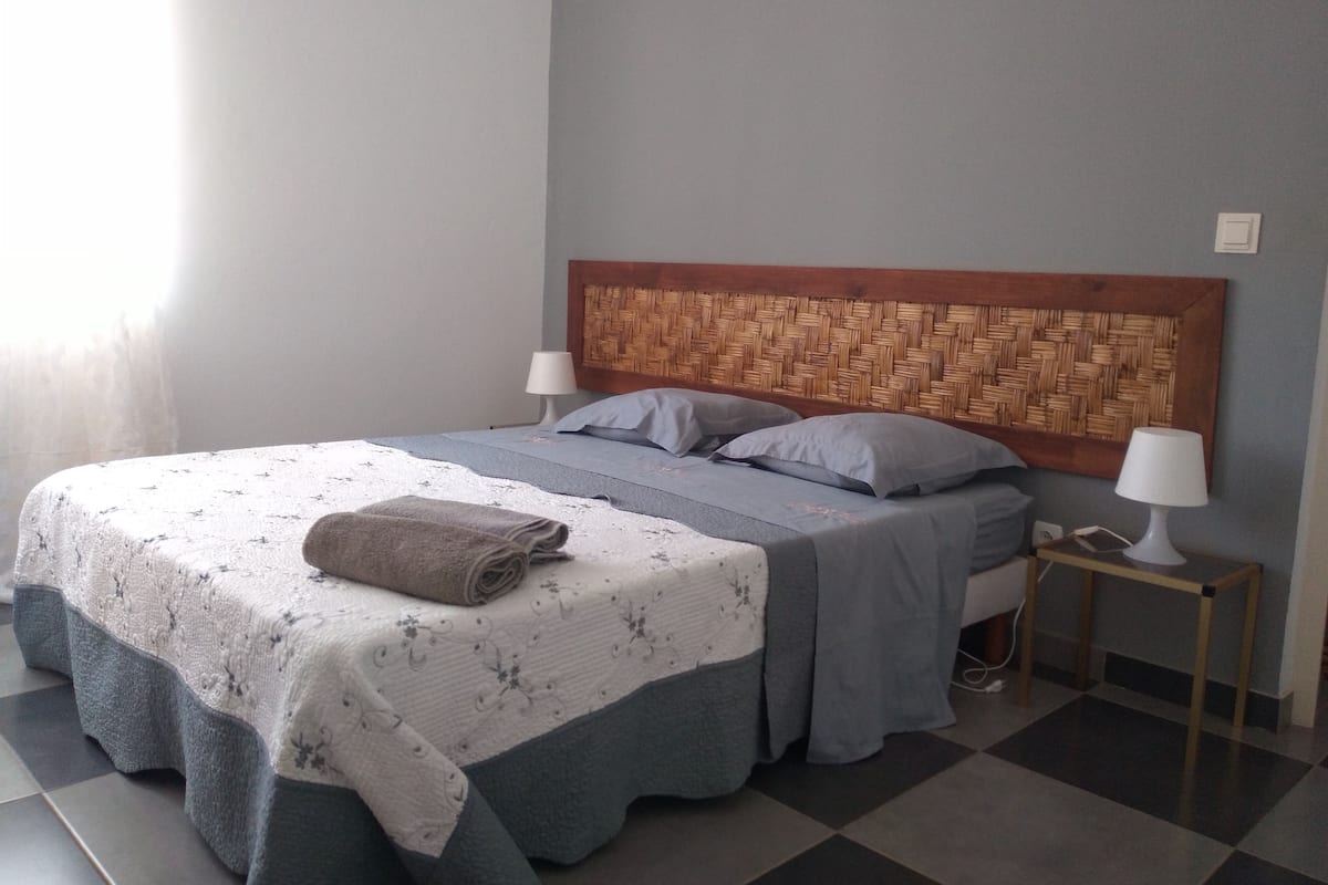Ein ordentlich gemachtes Bett mit einer gemusterten Bettdecke, zwei Kissen und einem aufgerollten Handtuch an der Seite. Am Kopfteil befindet sich ein Holzpaneel mit einem gewobenen Muster, ein Nachttisch mit einer Lampe und ein Schichtbodenbelag mit Schachbrettmuster.