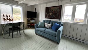A living room with a blue sofa, a television on a wooden stand, a painting on the wall, and a window with a view of the outside. - Oceanside Artists Cottage, Dunfield, Trinity Bay (Dunfield)