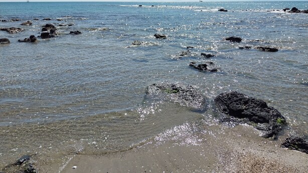Een strand met helder water, rotsen en verre zeilboten.