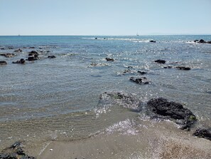 A beach with clear water, rocks, and distant sailboats.