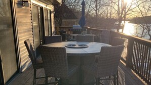 A wooden deck with a round table and four chairs, overlooking a body of water with a sunset in the background.