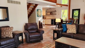 A spacious living room with a high ceiling, a wooden balcony, and a view of the kitchen.