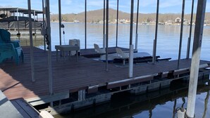 A covered dock with chairs and a table, overlooking a calm lake with boats and houses in the background.