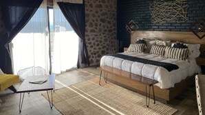 Un dormitorio con pared de piedra, techo de madera, cama con múltiples almohadas, una mesita de noche y una mesa con una silla.