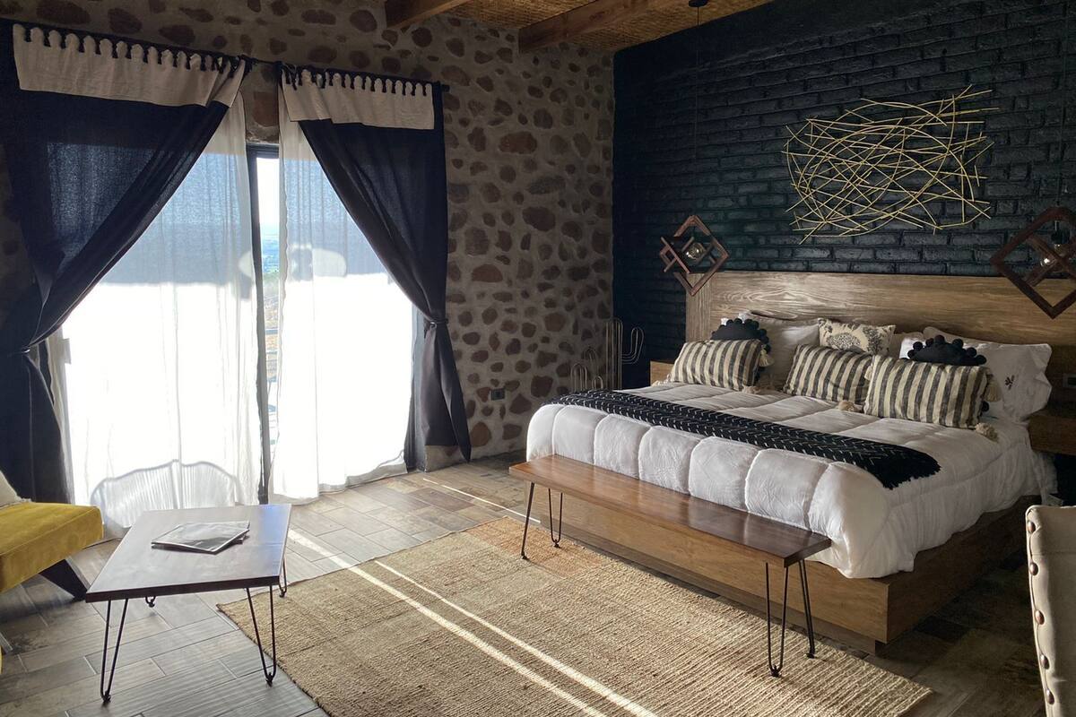 Un dormitorio con pared de piedra, techo de madera, cama con múltiples almohadas, una mesita de noche y una mesa con una silla.
