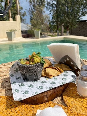 A table with a stone bowl of guacamole, tortilla chips, and a salt shaker by a poolside.