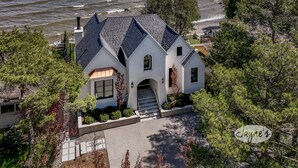 Une maison avec un toit à forte pente, de grandes fenêtres et des fondations en pierre, entourée d’arbres et donnant sur un plan d’eau.