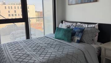 A bedroom with a large window, a bed with patterned bedding, a nightstand, and a painting on the wall.