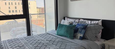 A bedroom with a large window, a bed with patterned bedding, a nightstand, and a painting on the wall.