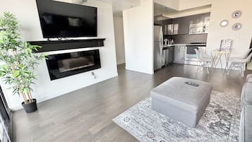 A modern living room with a flat-screen TV, a fireplace, a grey ottoman, and a dining area with a table and chairs.