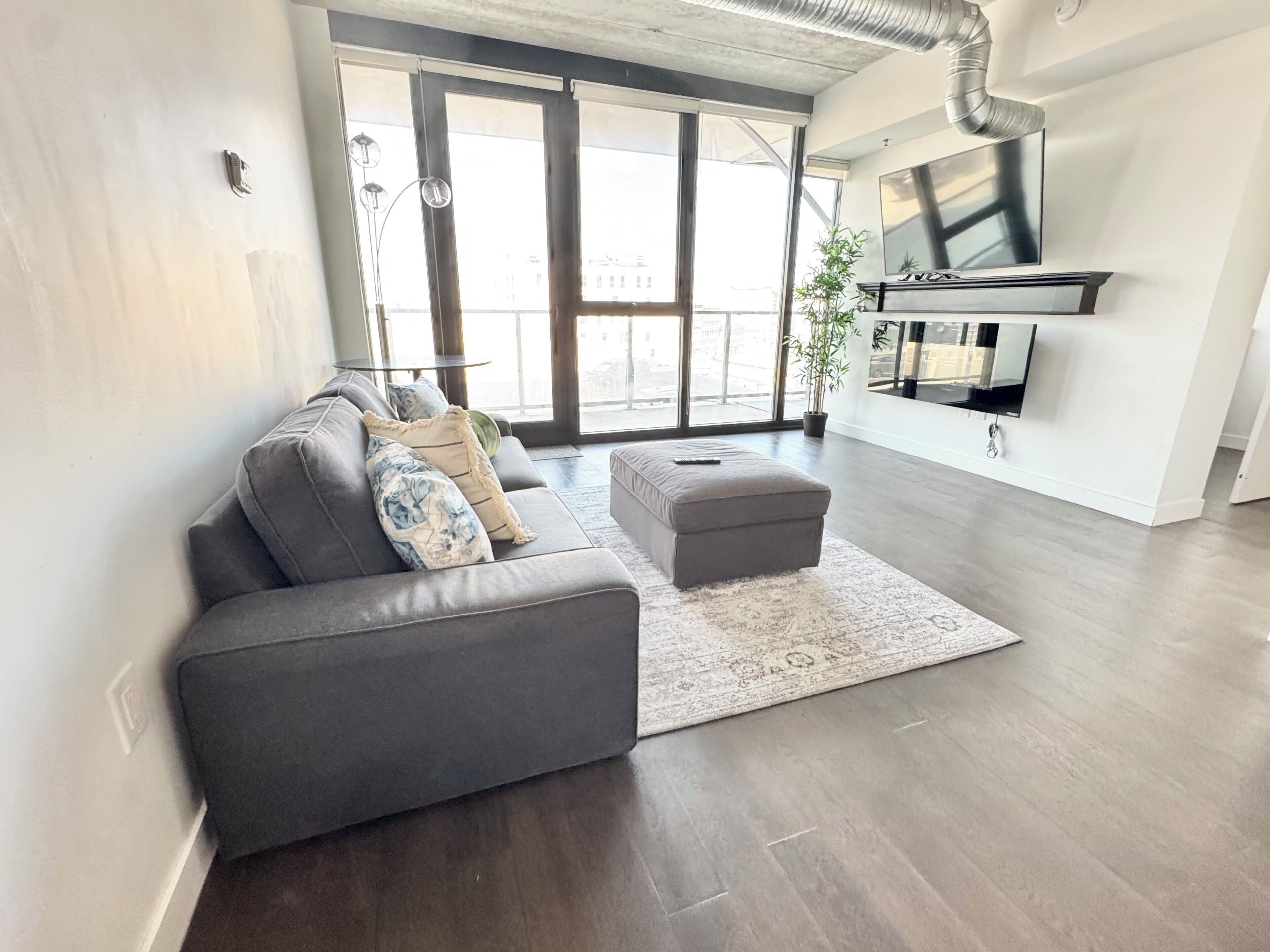A modern living room with a grey sofa, a matching ottoman, and a large window with a view of buildings.