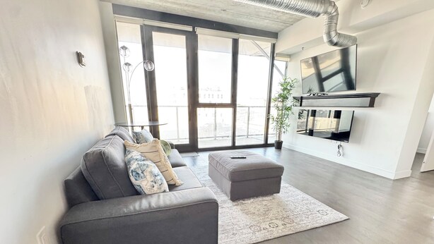 A modern living room with a grey sofa, a matching ottoman, and a large window with a view of buildings.