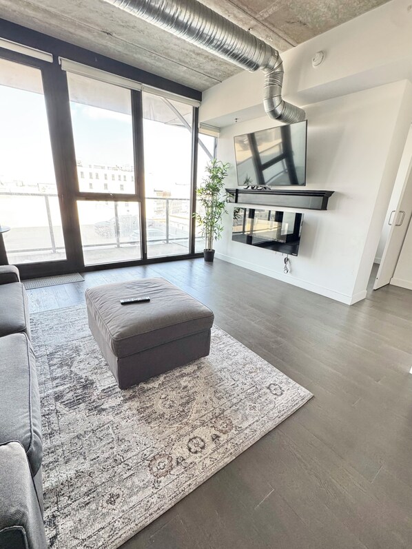A modern living room with a large window, a grey sofa, a patterned rug, and a flat-screen TV mounted on the wall. - Downtown across from arena wifi and laundry (Winnipeg)