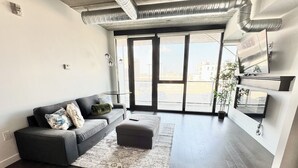 A modern living room with a grey sofa, a matching ottoman, and a large window with a view of buildings.