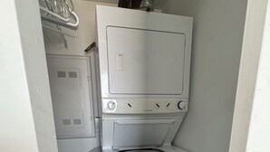 A compact laundry room with a front-loading washer and dryer, a small shelf above the dryer, and a metal rack with clothes.