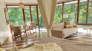 A bedroom with a large bed, a sofa, a dining table, and chairs. There is a view of trees through the windows. - 1960 Mid-Century Modern Nature Retreat (Cambridge)