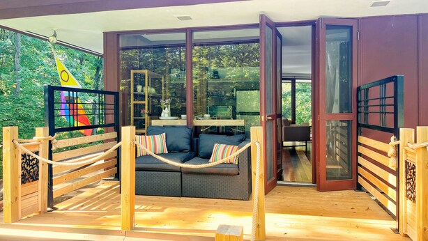 A modern outdoor seating area with a sofa and cushions, a wooden deck, and a glass door leading to an interior space. - 1960 Mid-Century Modern Nature Retreat (Cambridge)