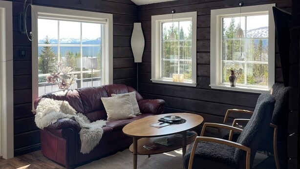A cozy room with a dark wooden interior, a leather sofa, a round coffee table, and two armchairs. Large windows offer views of a mountainous landscape. - Experience Fjällkroken, an Exquisite Mountain Cabin Built in 2016 (Undersåker)