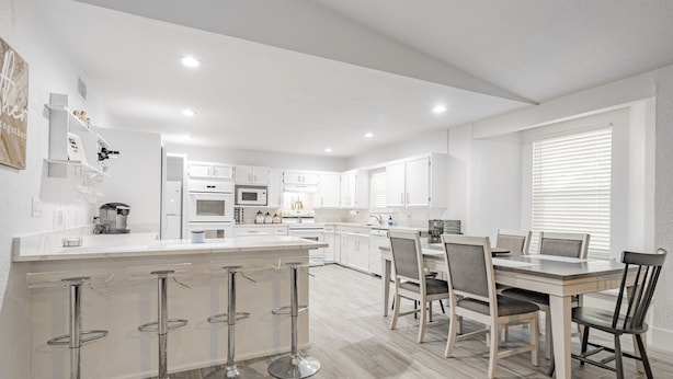 A modern kitchen with white cabinets, a central island with bar stools, and a dining area with a table and chairs.