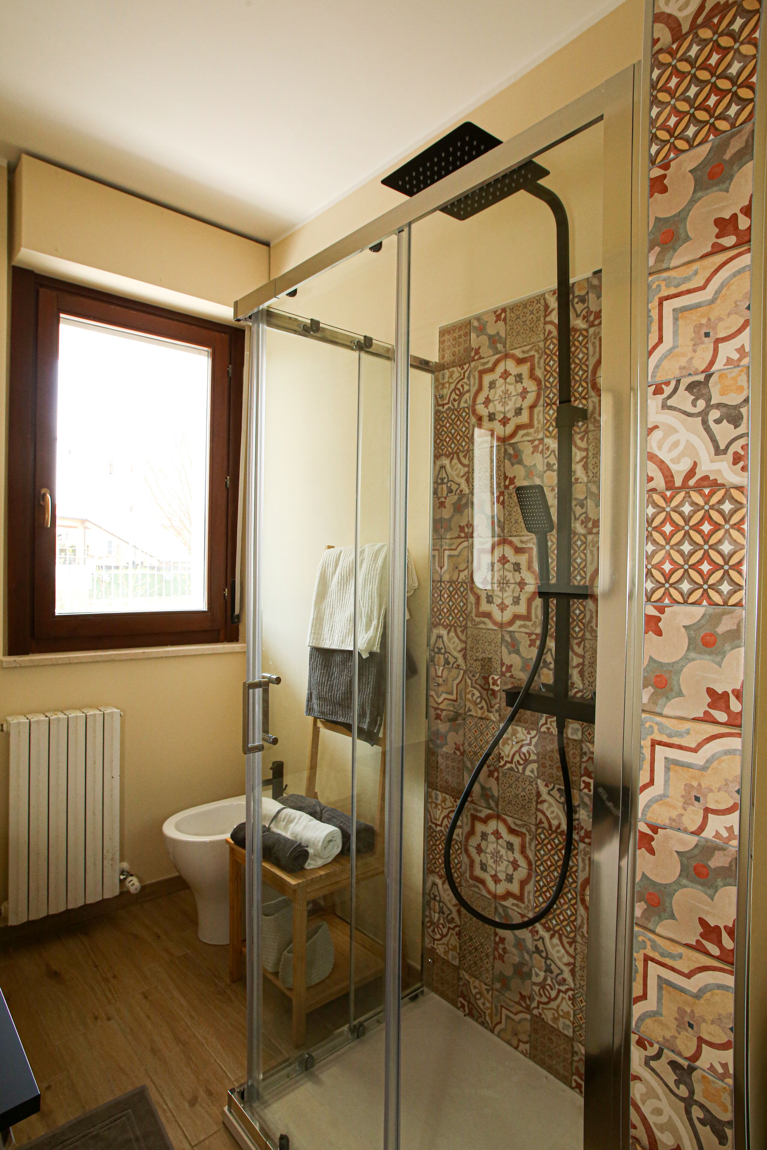 Un baño con cabina de ducha de vidrio, inodoro, ventana y baldosas estampadas.