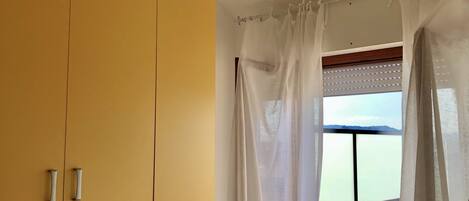 A room with a bed, yellow wardrobe, and a window with sheer curtains.