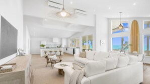 Uma sala de estar ampla com um sofá branco grande, uma mesa de centro e uma mesa de jantar com cadeiras. Uma cozinha aparece ao fundo, com vista para o oceano através de grandes janelas.
