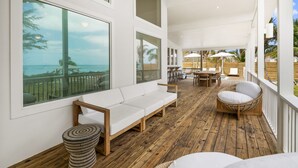 Uma área externa ampla com deck de madeira, mobiliário branco e grandes janelas com vista para o oceano.