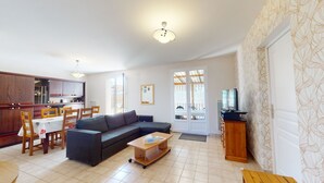 A living room with a sofa, coffee table, and TV stand. There is a dining area with chairs and a table. The room has a kitchen with cabinets and a window. - Dolce Vita close to the beach - Terrace & Garden (Saint-Hilaire-de-Riez)